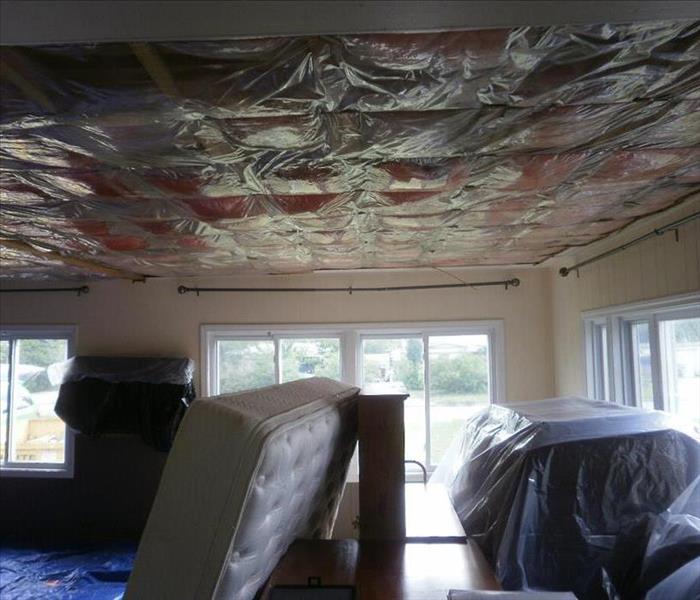 a living room with covered furniture, a mattress and plastic containment in ceiling where you can see pink insulation