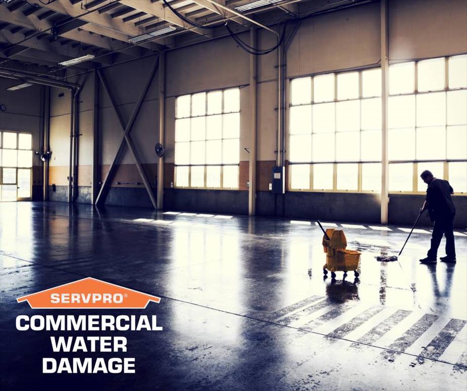 A big warehouse with water on floor and a man cleaning with a mop. SERVPRO logo and text commercial water damage below it